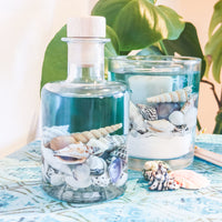 The blue sea reed diffuser is placed in front of the scented soy and gel  tumbler on a matching turquoise table top. Shells are scattered around and a large plant is in the background.